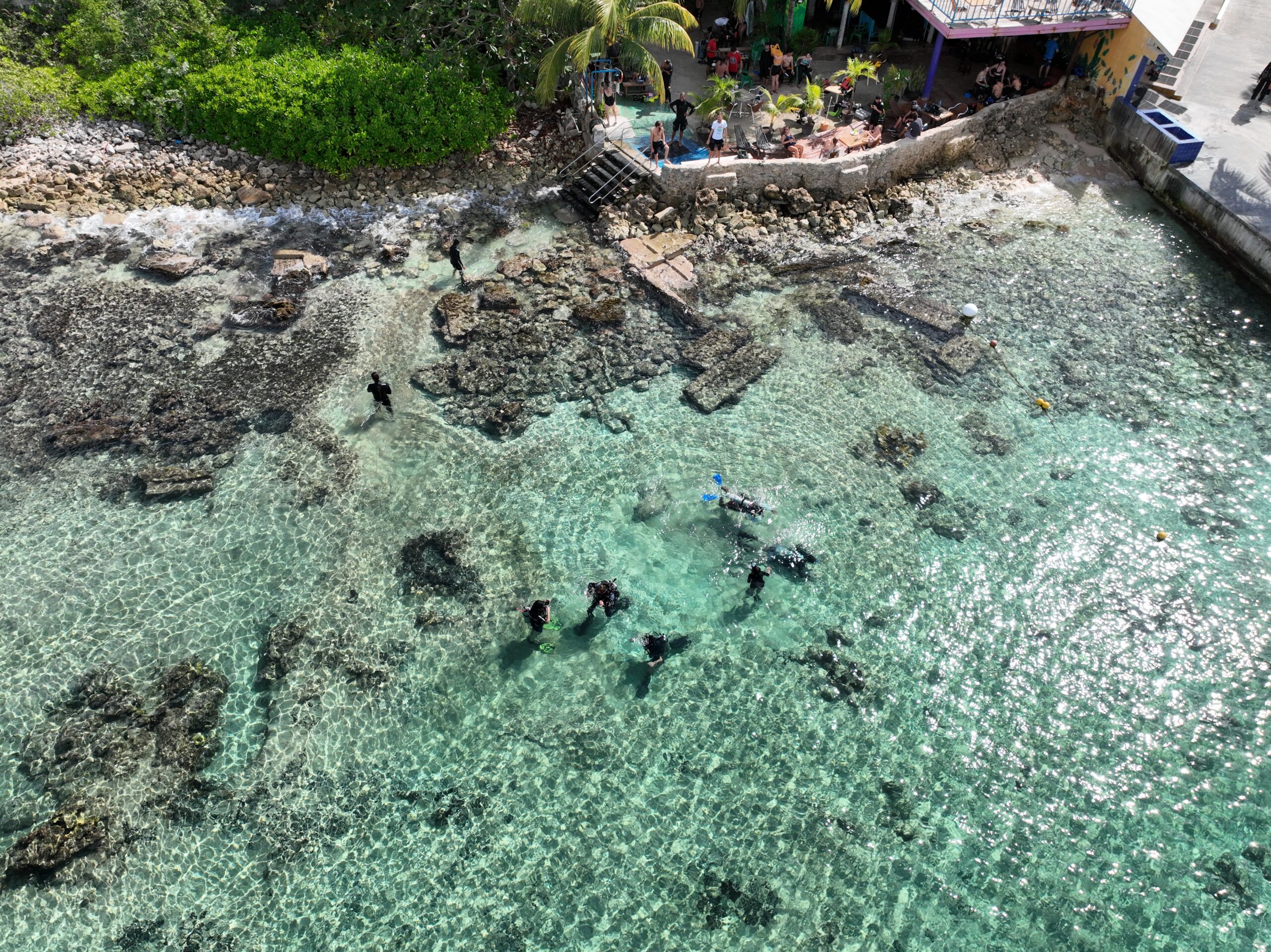 a view of divers practicing in the water at Tikila Beach in Cozumel, Mexico. The water in the photo is clear and turquoise in color, with several divers visible, wearing wetsuits and scuba gear. The divers could be seen practicing their skills, such as swimming, diving, and possibly exploring the underwater environment. The background show the sandy beach, palm trees, and other boats, piers, or structures in the surrounding area.