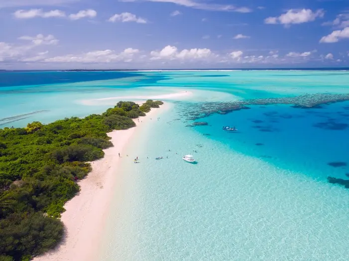 Aerial vue of the end of an island with different colors of cristal blue water, white sandy beach and trees. 2 boats and tourists can be seen bathing.