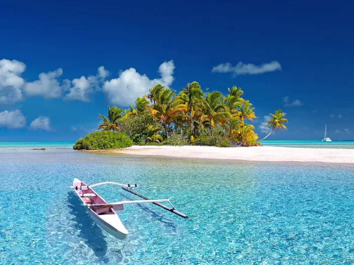 Wonderful vue of an island with palm trees and sandy white beach from the cristal clear water. There is a canoe in the first plan and a catamaran in the background.