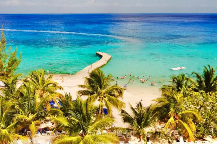 aerial view of Cozumel beach with palm trees in first plan followed by a view of white sandy beach, a stone pier and to finish different blue shades of cristalin water in the background