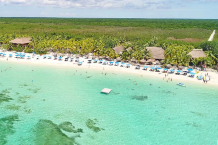 aerial vue of a Cozumel Beach Club with cristallin turquoise water in the first plan followed by blue beach unbrellas and seats on the beach, a few thatch roof buildngs, Palm trees and jungle in the background