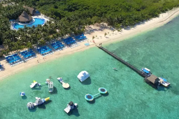 aerial view of Cozumel Paradise Beach Club, with water inflatable games, a wooden pier with a roof palapa at the end, 2 boats on the other end. Blue unmbrellas and beach chairs are set up on the beach, followed by a forest of palm trees with a swimming pool in the middle of the forest.