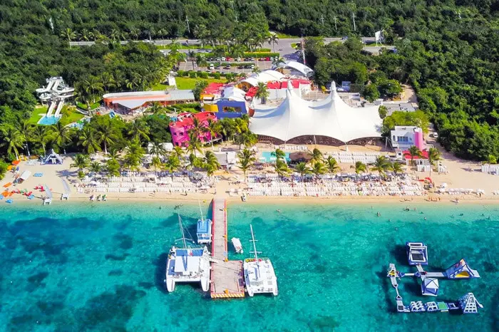 Aerial view of Playa Mia Beach Club, cristallin water supporting a concrete pier with catamarans and boats tied up to it. On the right there are inflatable games on the same shallow cristallin water. the beach is cover by lounge chairs and palm trees